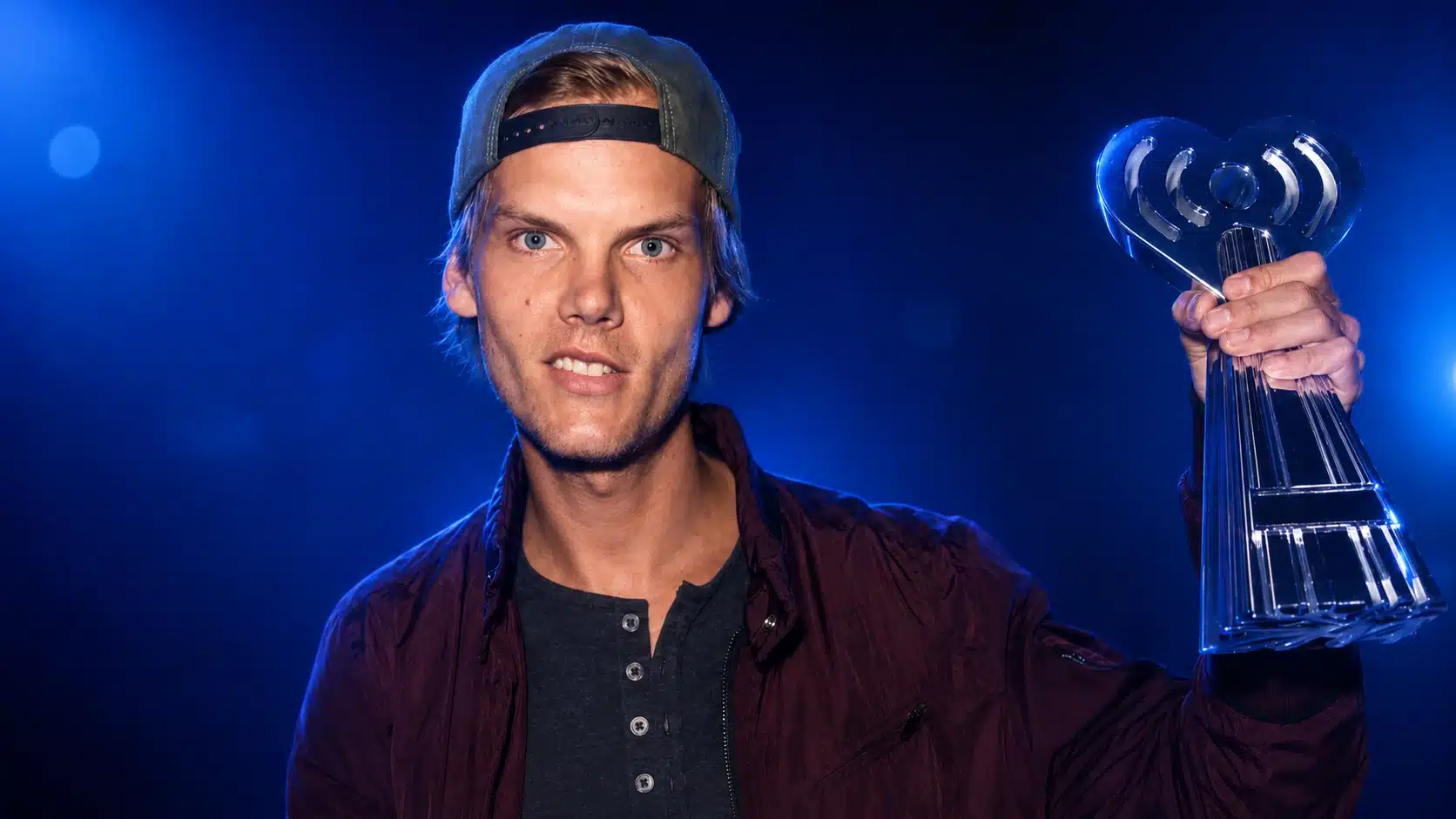 young man in burgundy jacket and backward cap holds clear trophy against dramatic blue stage lighting