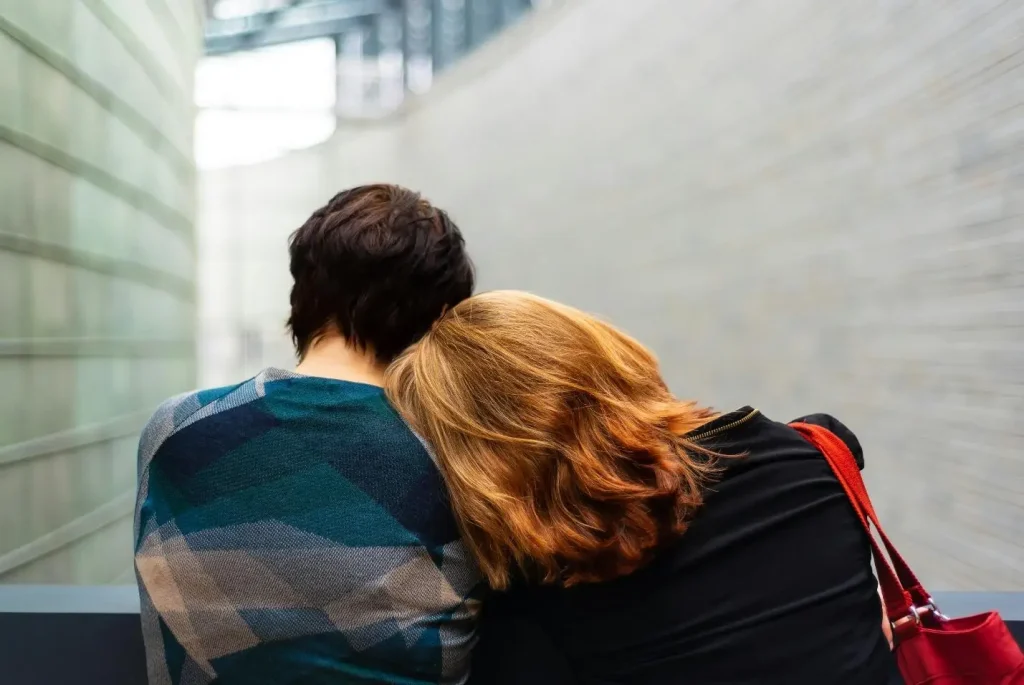 Two people sitting closely with one resting head on the other's shoulder in a modern building