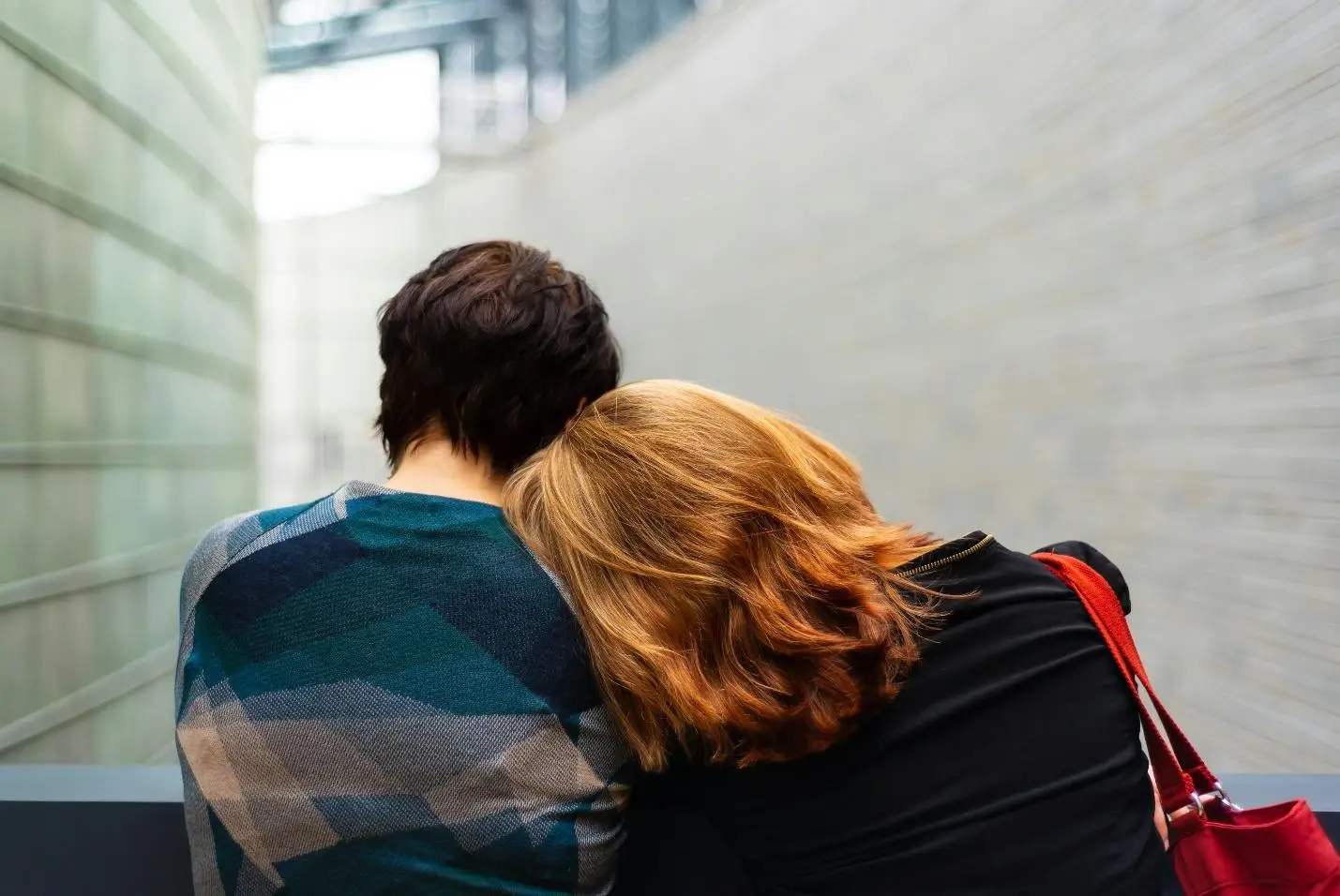 Two people sitting closely with one resting head on the other's shoulder in a modern building
