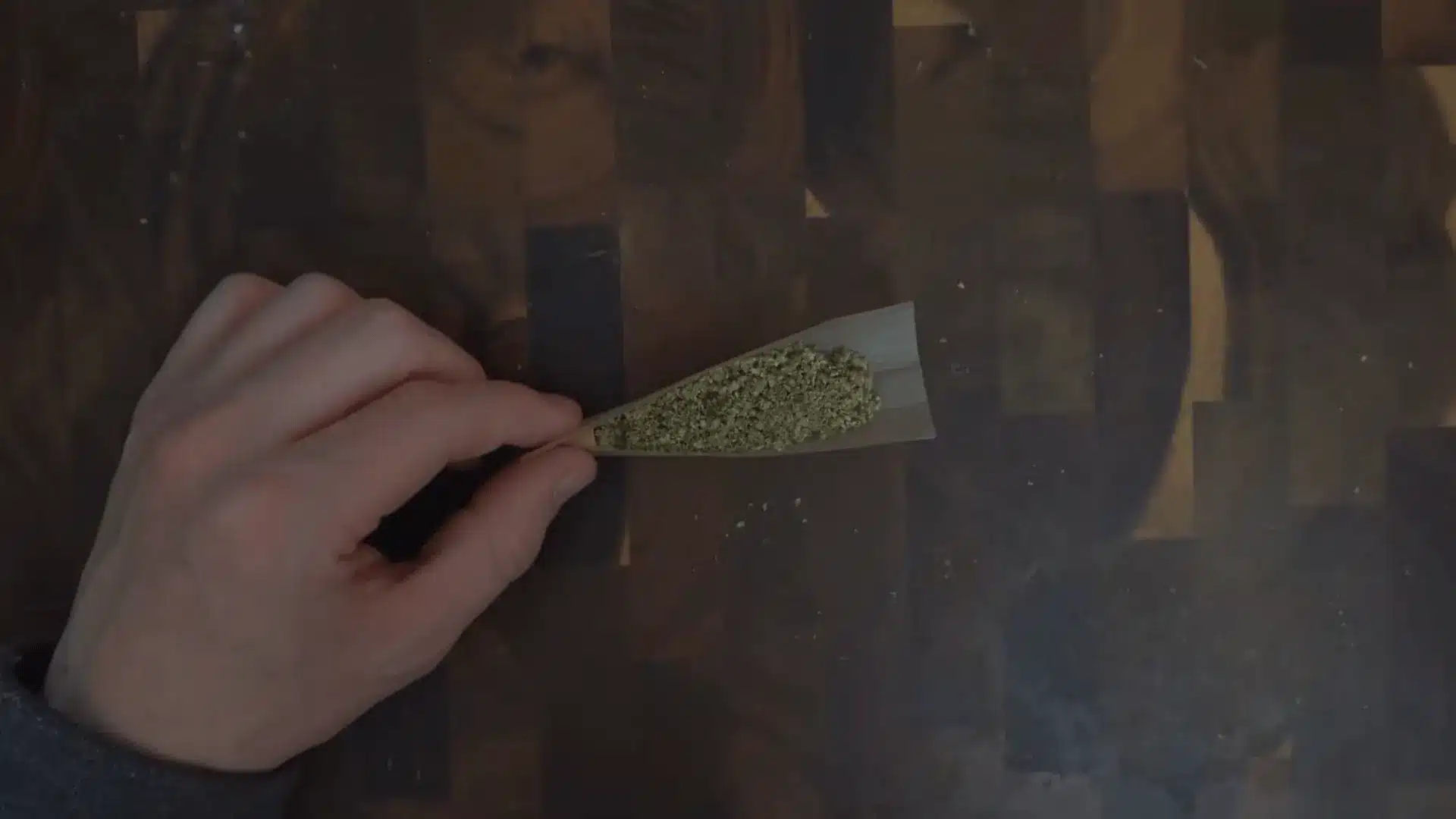 a hand holds an open rolling paper filled with ground cannabis above a dark wooden tabletop