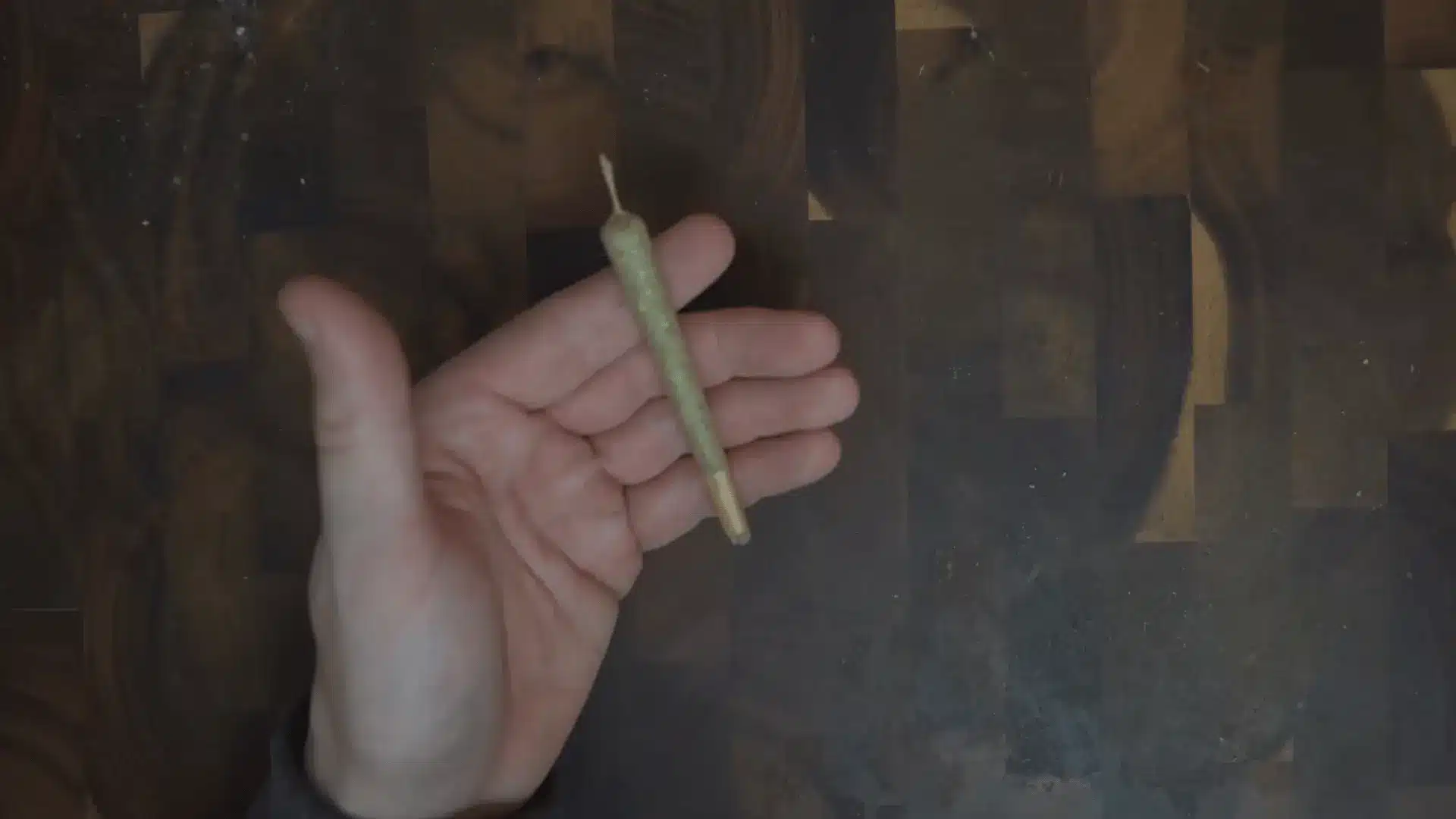 a hand displays a finished cannabis joint with twisted paper at one end over a dark wooden tabletop