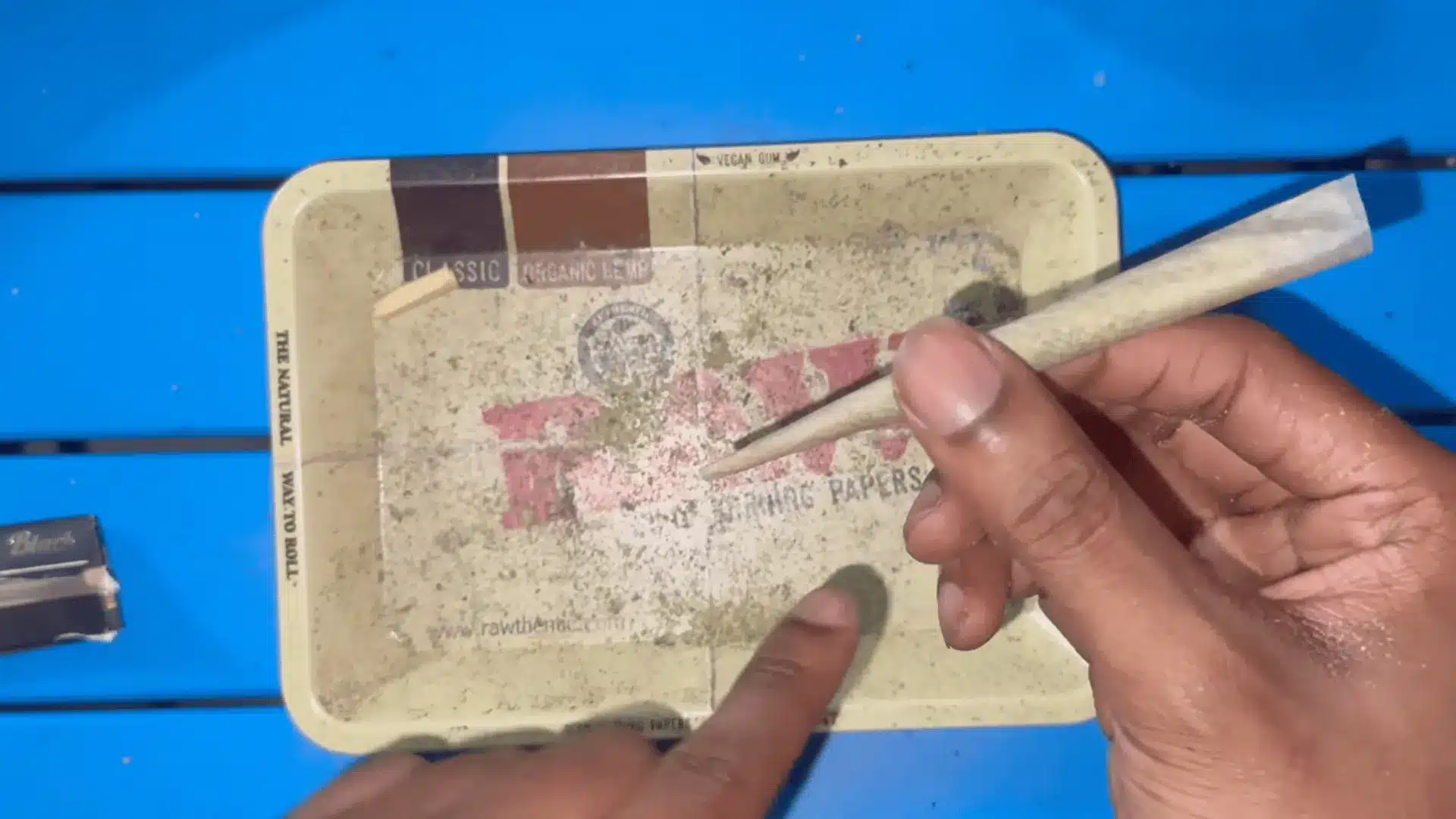 a hand holds a finished rolled paper above a worn tray while another finger points at loose herbs