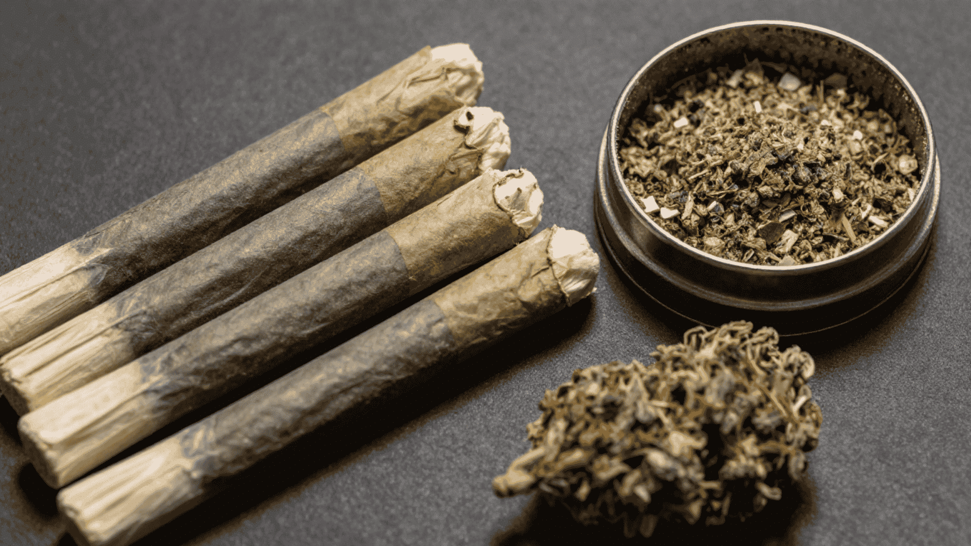 four cannabis joints beside a grinder filled with dried herbs and a loose cannabis bud