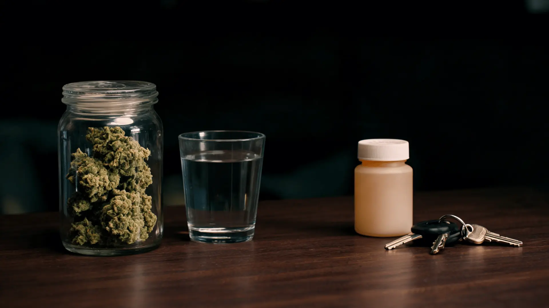 glass jar filled with cannabis buds beside a glass of water a pill bottle and car keys on a wooden table against a dark background