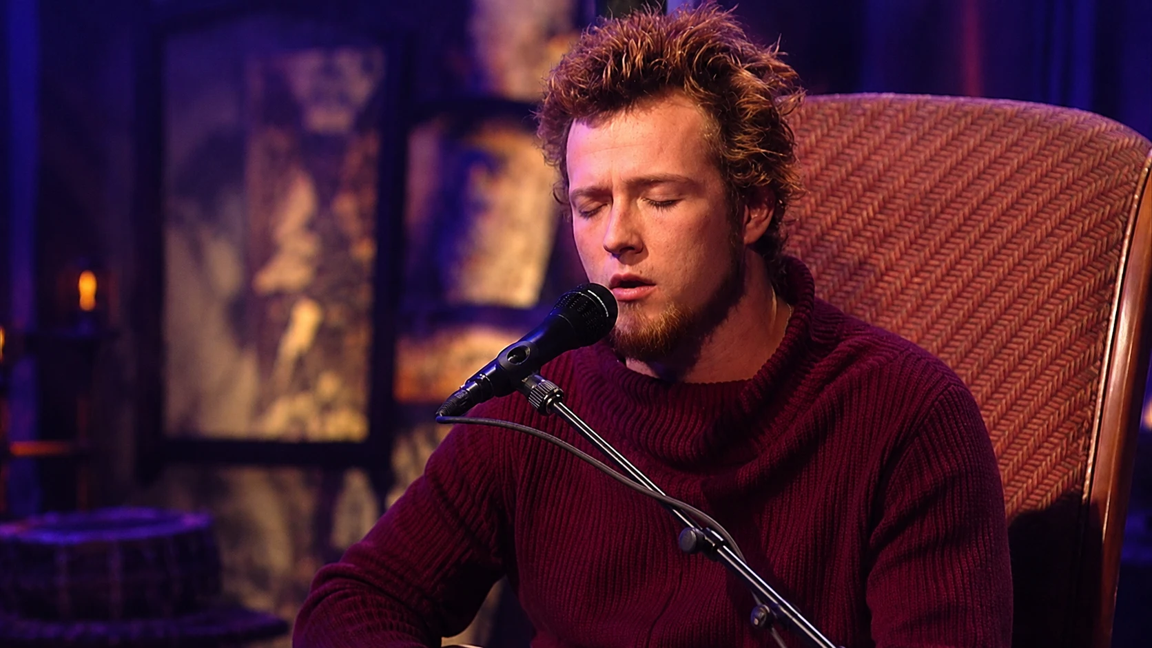 male singer performing into a microphone on stage, wearing a burgundy sweater under warm purple and amber lighting
