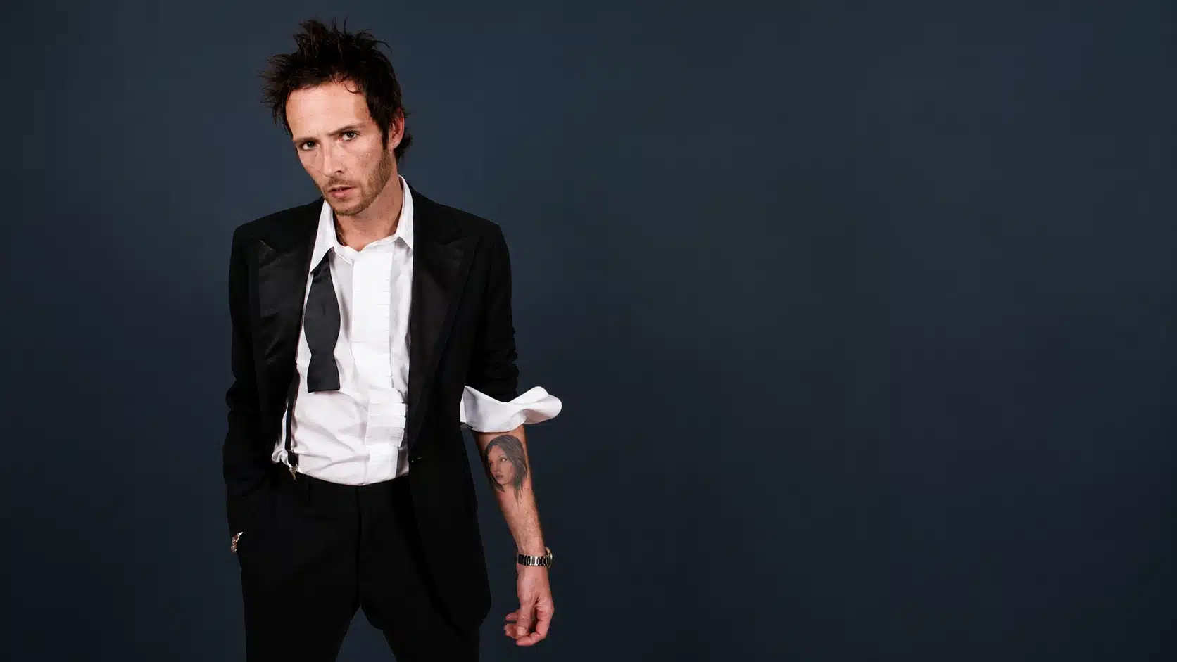 man in black suit with loose bow tie poses against dark blue studio background showing forearm tattoo