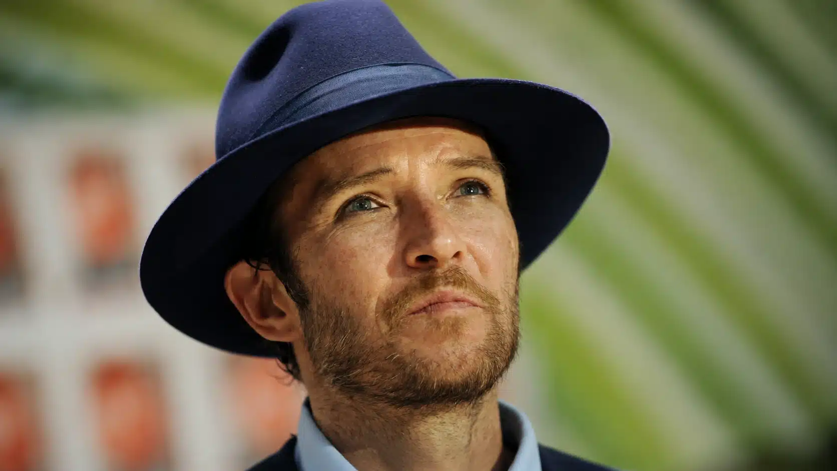 man wearing a navy fedora looks upward in a close-up portrait with soft green background and natural lighting