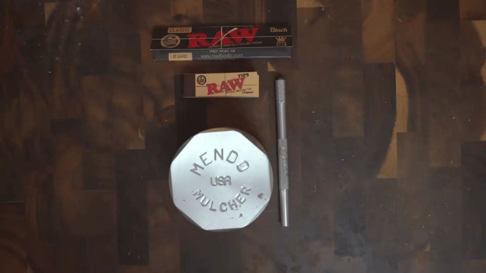 rolling papers tips a metal tool and a white grinder arranged on a dark wooden tabletop from above