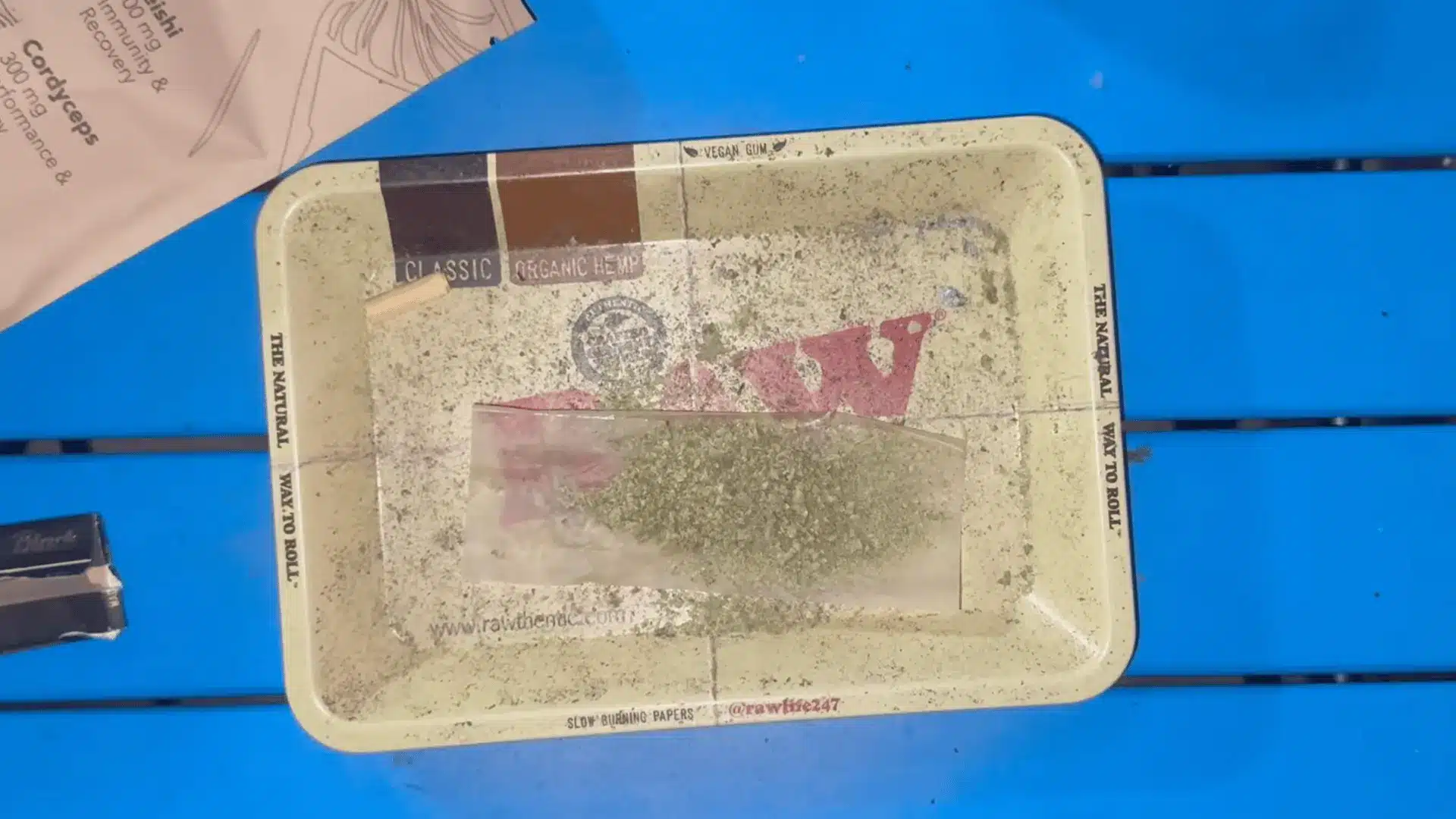 ground herbs rest on a rolling paper inside a worn tray placed on a bright blue slatted table