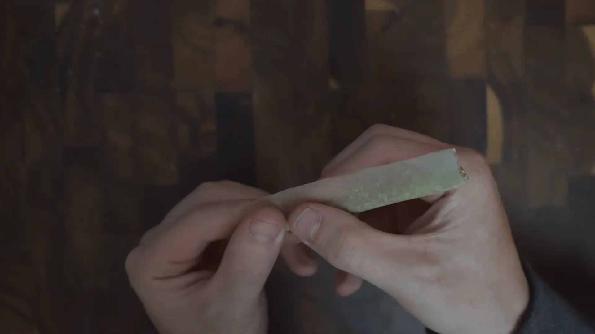 two hands roll a cannabis filled paper into a narrow cylinder above a dark wooden tabletop