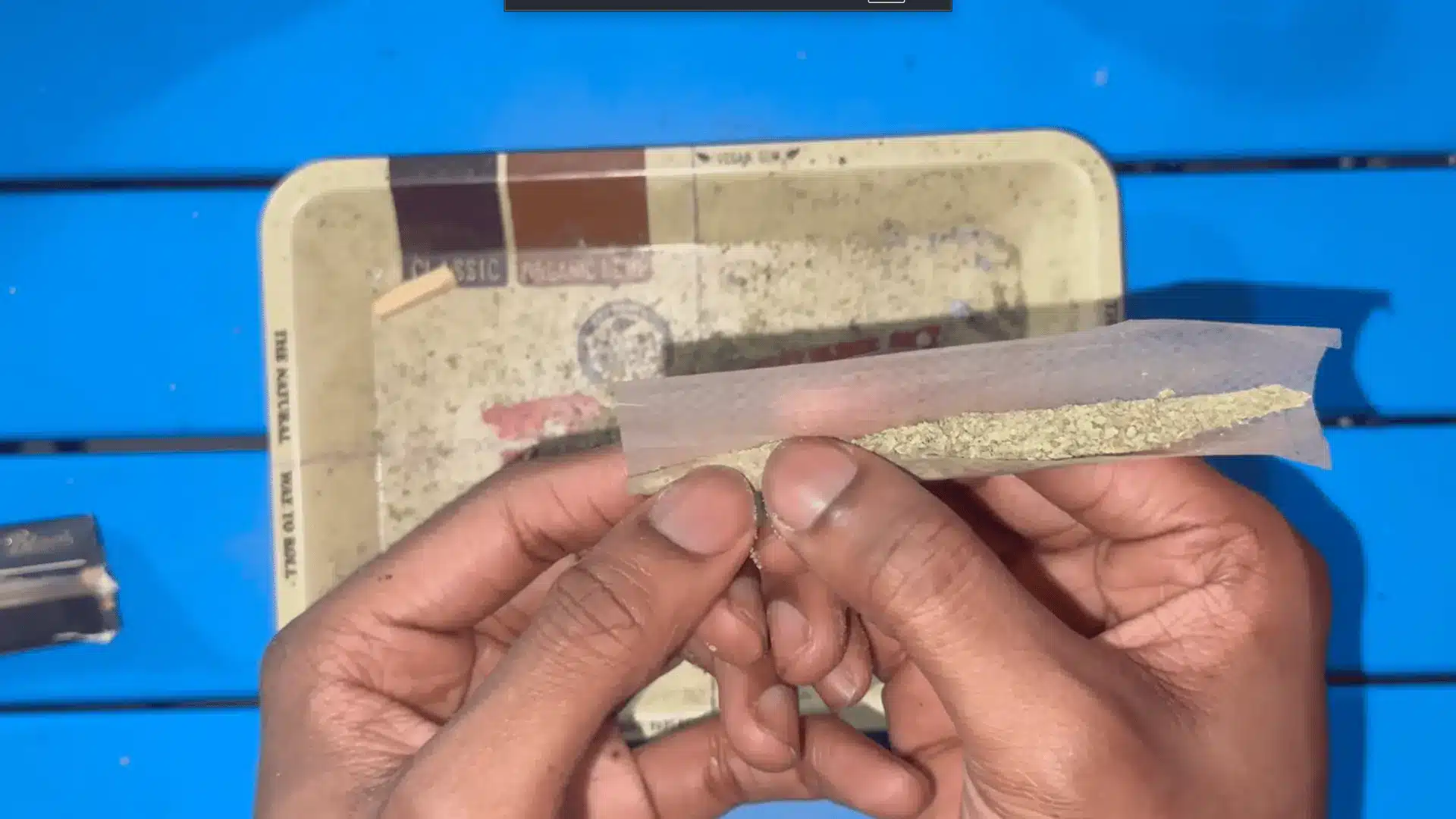 two hands hold a rolling paper filled with ground herbs above a worn tray on a bright blue table