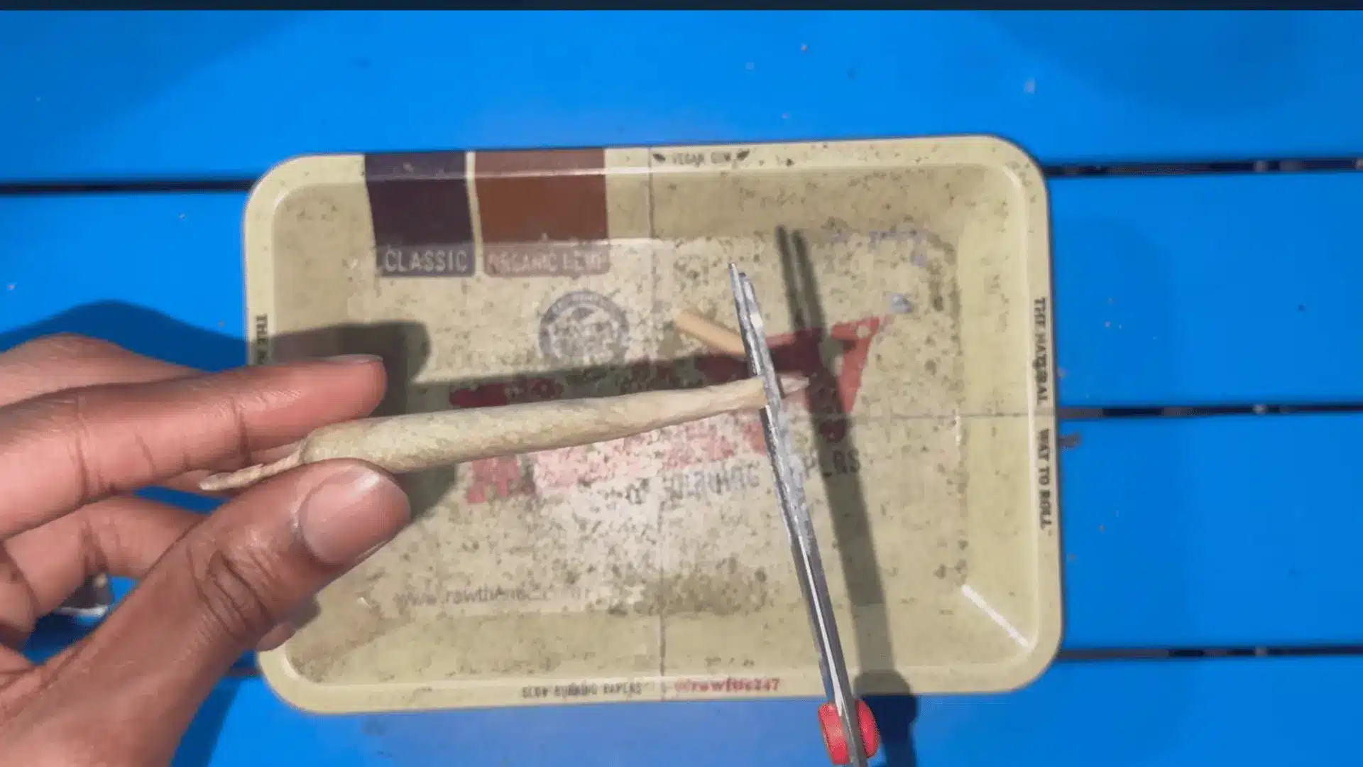 a hand holds a rolled paper while scissors trim one end above a worn tray on a bright blue table